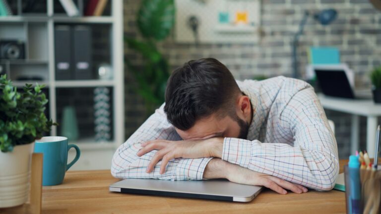 A man laying his head down over his folded arms on a desk.